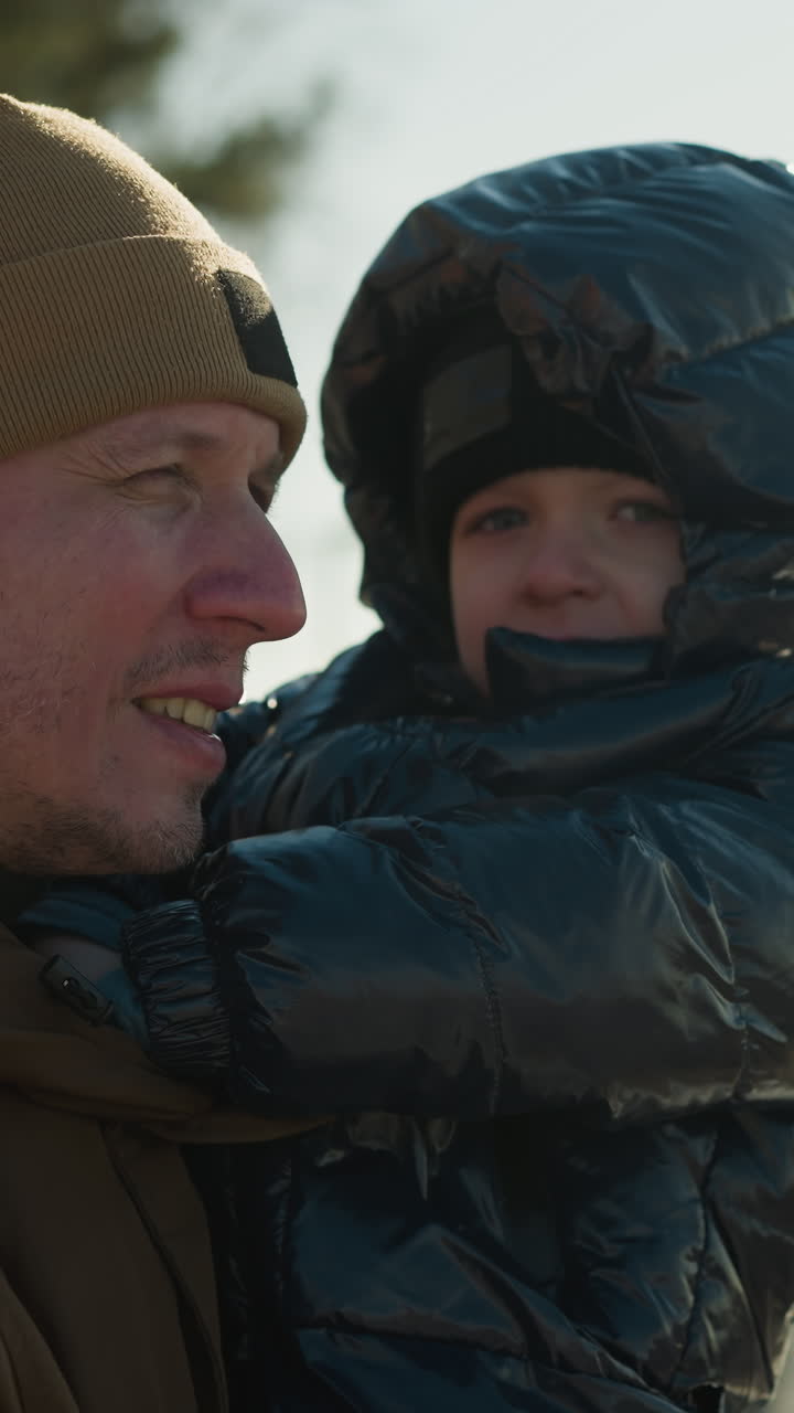 A father stands close to a metal railing, carrying son in his arms, the father gazes over a cityscape while his son looks directly at the camera, both dressed warmly in jackets and beanies