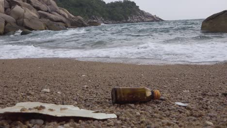 Littered-glass-bottle-and-plastic-foam-left-on-a-beach-near-wave,-zoom-out-shot