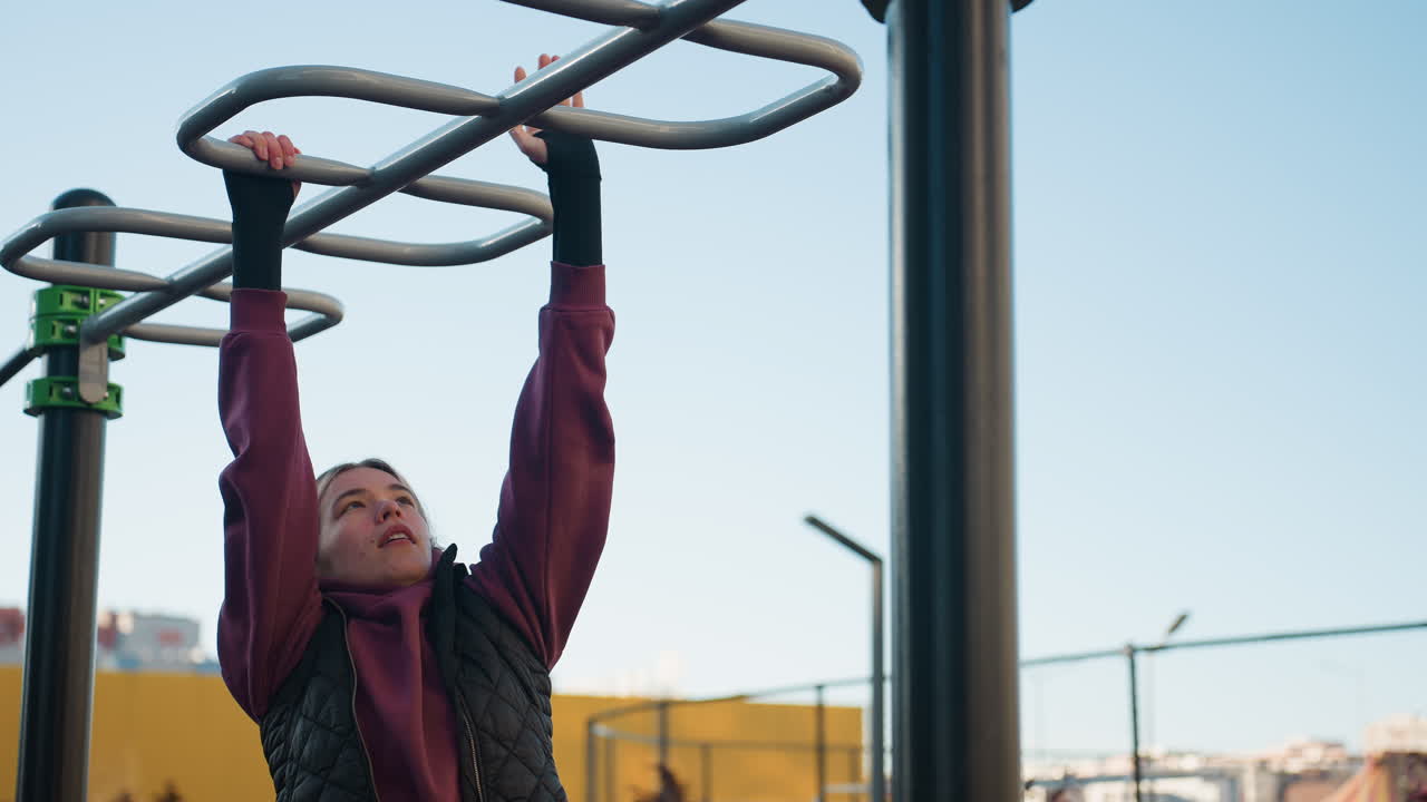 Female athlete in maroon hoodie hangs from silver pull up bars in outdoor park under clear sky, focus visible on her face, dynamic calisthenics showcasing strength, balance, resilience