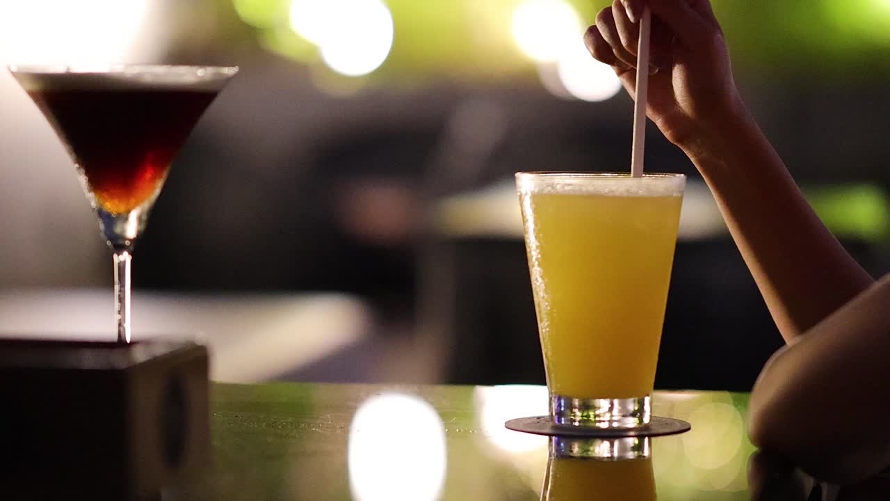 Person stirs yellow-green cocktail with straw beside martini glass in softly lit lounge