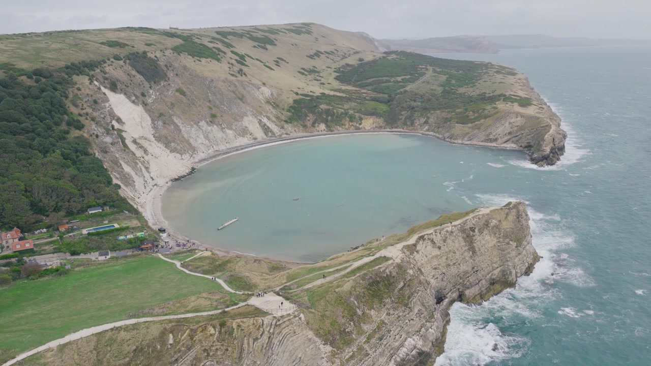 Veduta aerea dall'alto di Lulworth Cove nel Dorset che mostra l'intera baia curva, le scogliere calcaree, i sentieri costieri, la spiaggia e il promontorio circostante della Jurassic Coast
