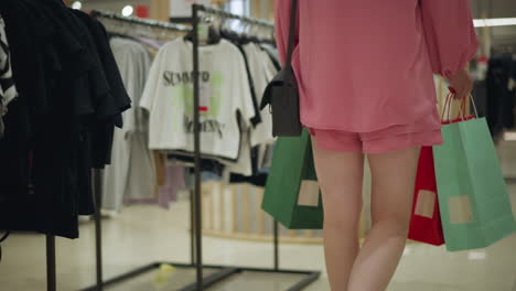 white lady in pink outfit and canvas shoes walking through a well-lit shopping mall, she carries a shopping bag, passing by neatly arranged clothes in different sections of the store