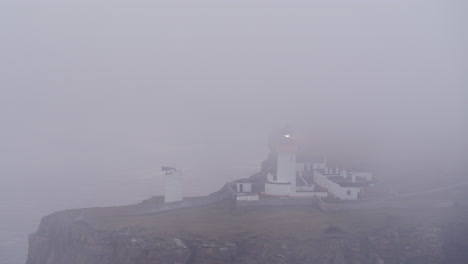 Luftaufnahme-Eines-In-Nebel-Gehüllten-Leuchtturms,-Dessen-Licht-Durch-Die-Dunkelheit-Strahlt