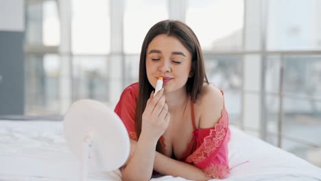 Beautiful-girl-applies-hygienic-lipstick-looking-in-the-mirror-while-lying-on-the-bed