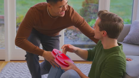 man surprising same sex partner at home with heart shaped gift held behind his back