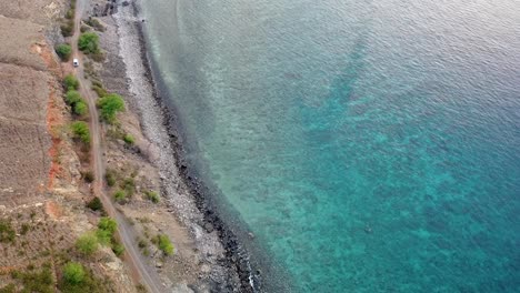 Luftdrohne-Eines-Weißen-Autos,-Das-Sich-Langsam-Entlang-Einer-Rauen,-Holprigen-Küstenstraße-Mit-Türkisfarbenem-Ozean-In-Timor-Leste-Bewegt