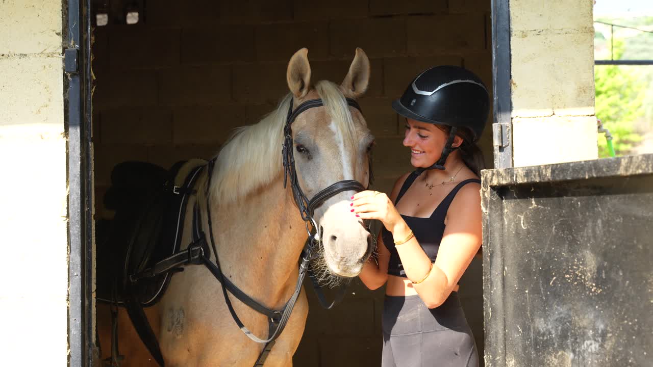 Smiling woman gently pets her horse in the stall as she talks with someone nearby