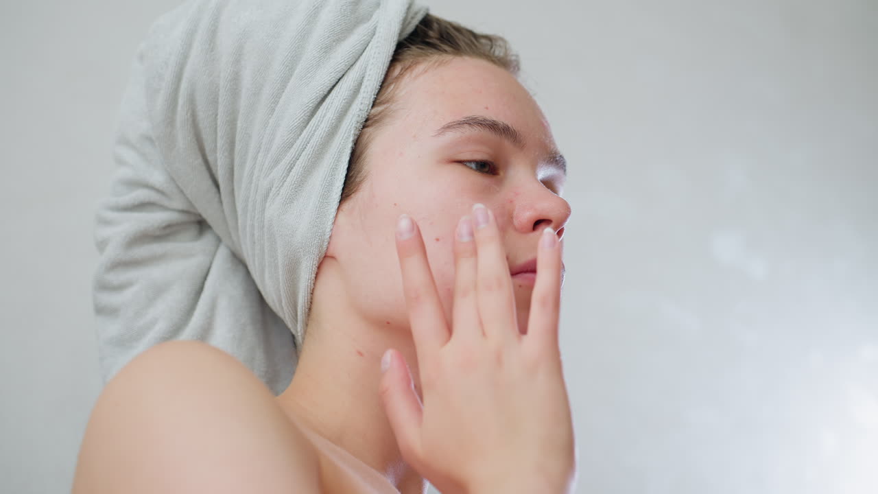 Closeup view of young girl with eyes closed gently applying skincare cream to cheeks with fingertips, towel wrapped on head, engaging in moisturizing, facial care, personal wellness, beauty ritual