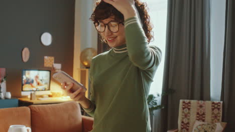 smiling asian girl in headphones dancing with phone in hand at home