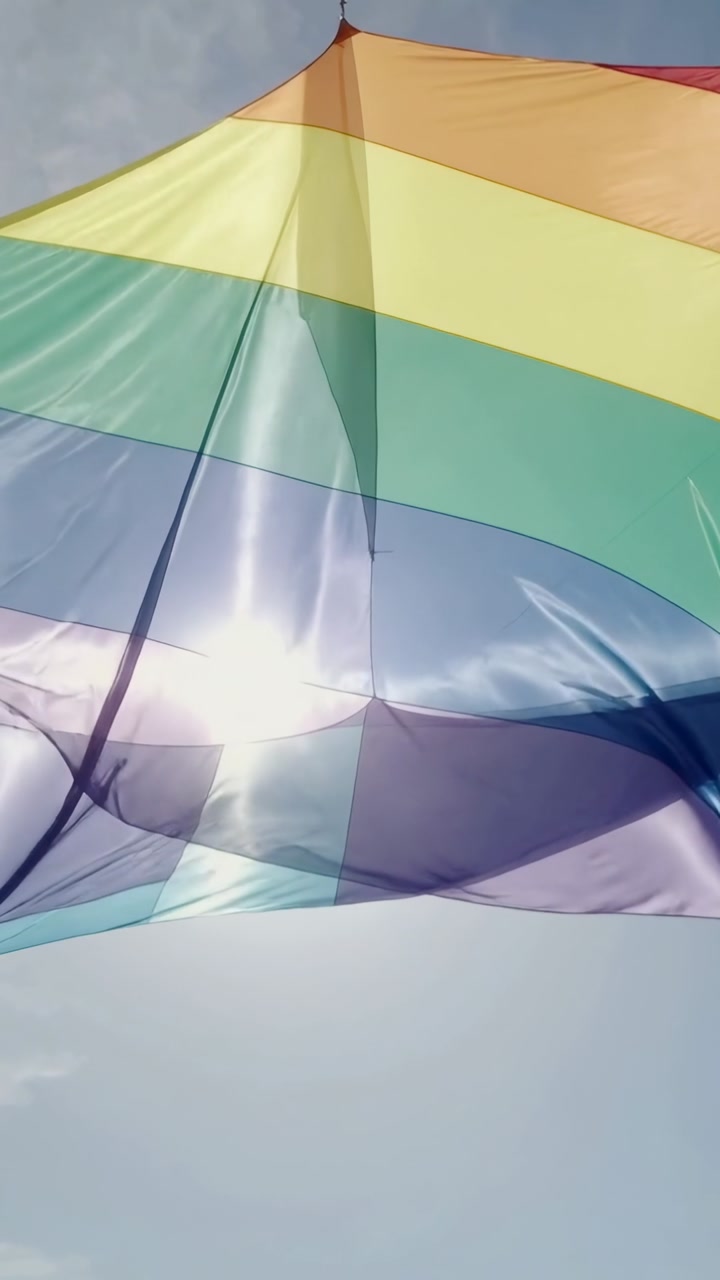 Vertical video: Billowing rainbow flag fluttering on breeze in open air, catching sun, pole visible
