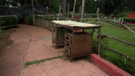 Un-Viejo-Escritorio-Rústico-Se-Encuentra-Al-Aire-Libre-En-Un-Exuberante-Parque-Verde,-Cerca-De-Un-Camino-Erosionado