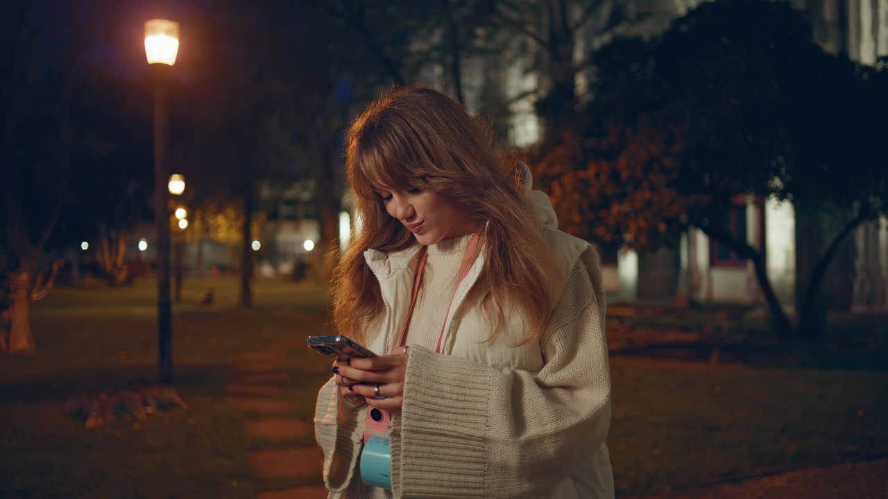 Donna sorridente che messaggia al telefono in una strada notturna illuminata dai lampioni, primo piano.