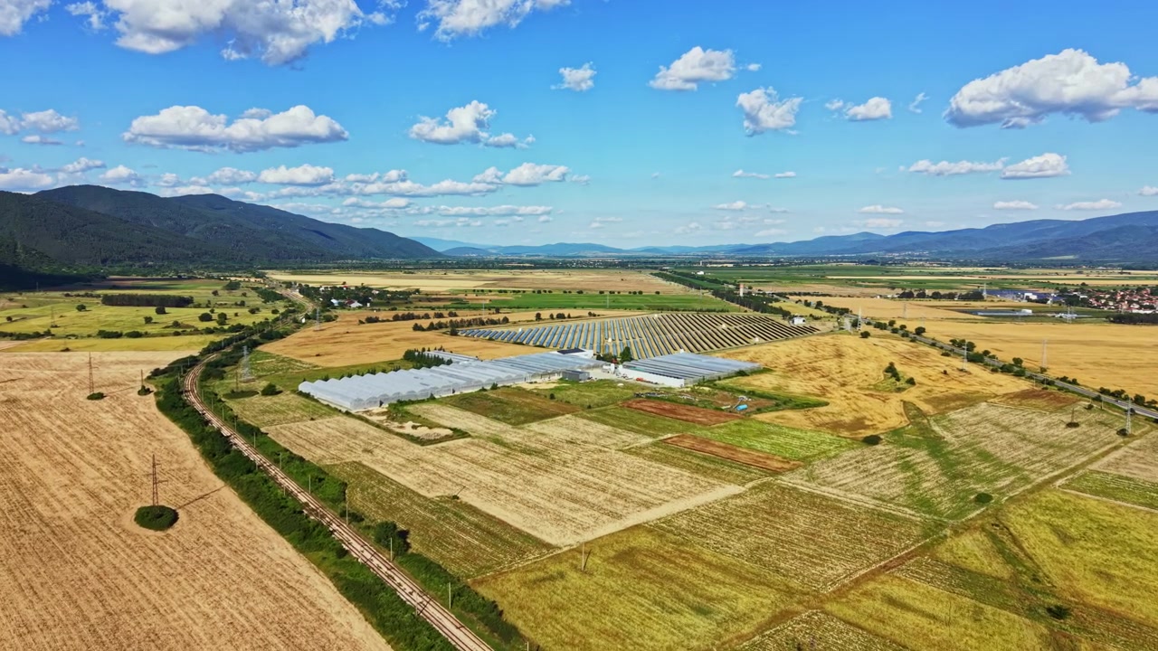 Weitwinkelaufnahme von landwirtschaftlichen Feldern und Gewächshäusern in einer ländlichen Gegend, mit Bergen und blauem Himmel im Hintergrund bei Tageslicht.
