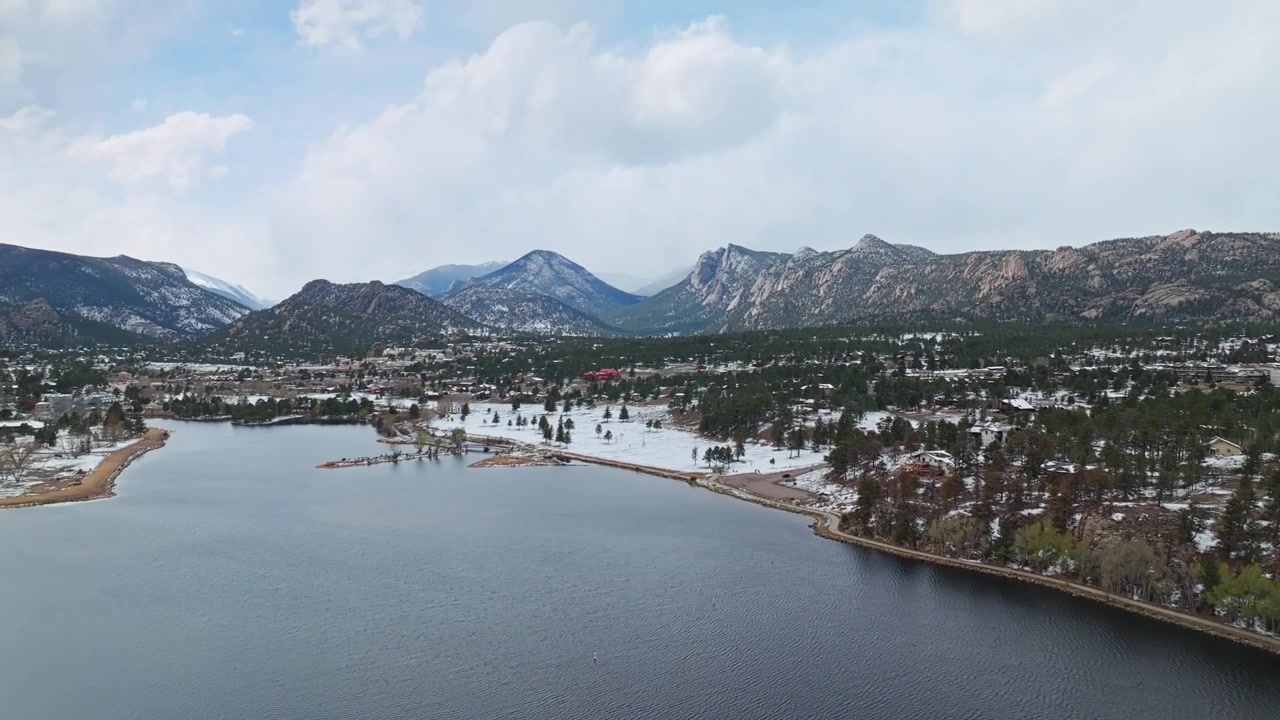Quang cảnh từ trên cao bờ hồ Estes Park với rừng cây và núi tuyết dưới bầu trời nhiều mây, quay bằng drone.