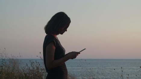 girl standing on a cliff by the sea on sunset- golden hour, with tablet computer in hands close