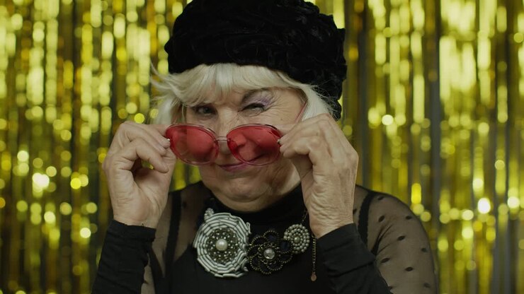 Adorabile anziana signora matura che tocca occhiali da sole rossi e ammicca giocosamente, flirtando e strizzando l'occhio