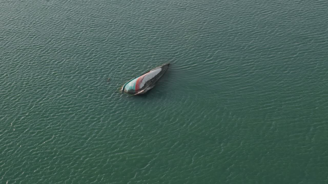 Ripresa aerea che si allontana all'indietro mostrando dall'alto una barca capovolta, per poi espandersi in un grandangolo che rivela la costa tropicale e le acque color smeraldo