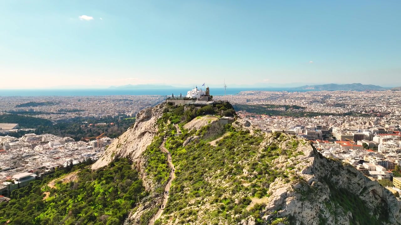 Veduta panoramica aerea della Collina del Licabetto e del paesaggio urbano di Atene sotto un cielo azzurro limpido.