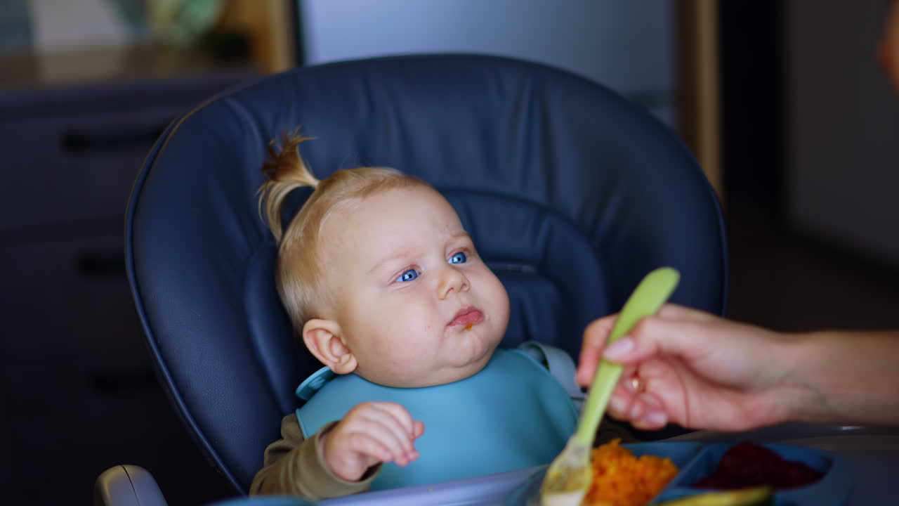 Blond kid in a blue bib sits at the high chair. Unrecognized mother feeds her child from a spoon.
