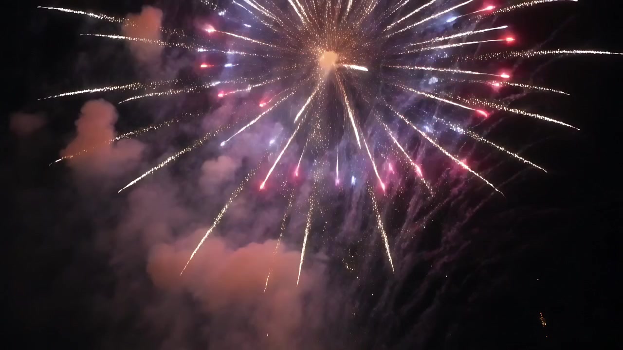Un fuoco d'artificio che esplode, inviando strisce radiali nel cielo notturno, con scintille che cadono e fumo.