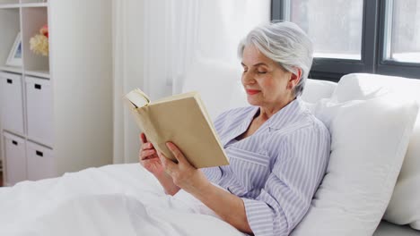 Mujer-Mayor-Leyendo-Un-Libro-En-La-Cama-En-El-Dormitorio-De-Su-Casa.-Tecnología,-Concepto-De-Vejez-Y-Personas.-Mujer-Mayor-Leyendo-Un-Libro-En-La-Cama-En-El-Dormitorio-De-Su-Casa.