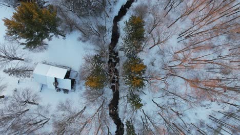 Top-view-of-a-beautiful-winter-landscape-with-snow-covered-forest-and-a-small-lonely-house-next-to-small-river