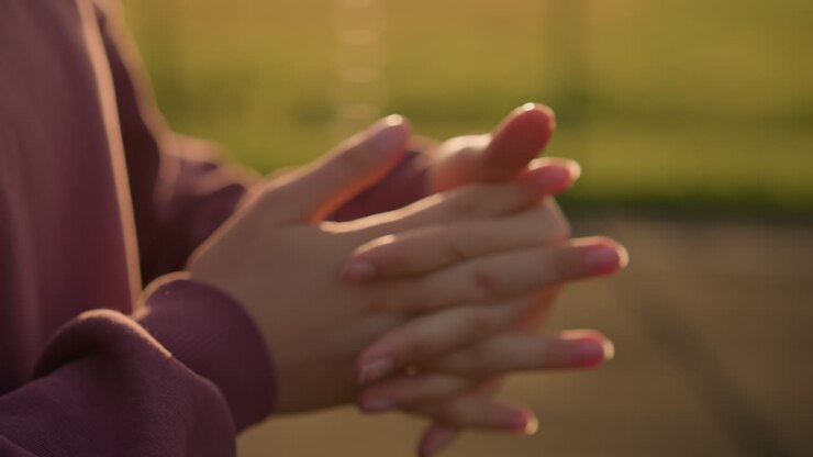 Primer plano de una mano de una persona vestida de color burdeos trabajando en la muñeca con los dedos entrelazados, con la luz del sol creando un efecto brillante y un fondo verde difuminado