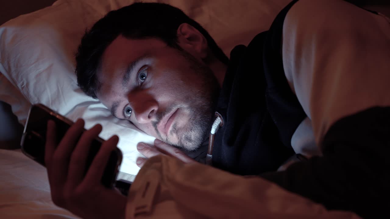 hombre acostado en la cama con un teléfono inteligente, la pantalla ilumina la cara en un dormitorio oscuro, desplazándose, redes sociales, enviando mensajes de texto a amigos en la noche antes de dormir