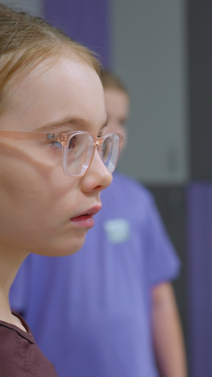 primer plano de una joven con gafas descansando en un poste mientras observa a otras dos niñas en el fondo, una vestida de rosa interactuando, la escena se desarrolla en un gimnasio o instalación de entrenamiento