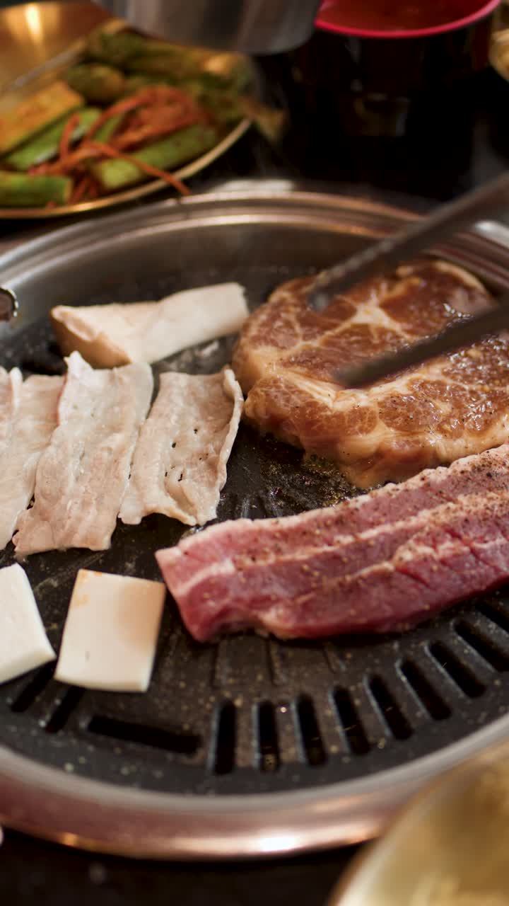 Daging babi mentah bagian perut dan steak tersusun di atas panggangan Korea meja dengan penjepit, pencahayaan dalam ruangan yang hangat.