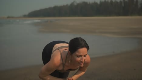 Chica-Practicando-Yoga-En-La-Playa-De-Arena-Negra-Haciendo-Pliegues-Hacia-Adelante-Y-Saltando-A-La-Tabla