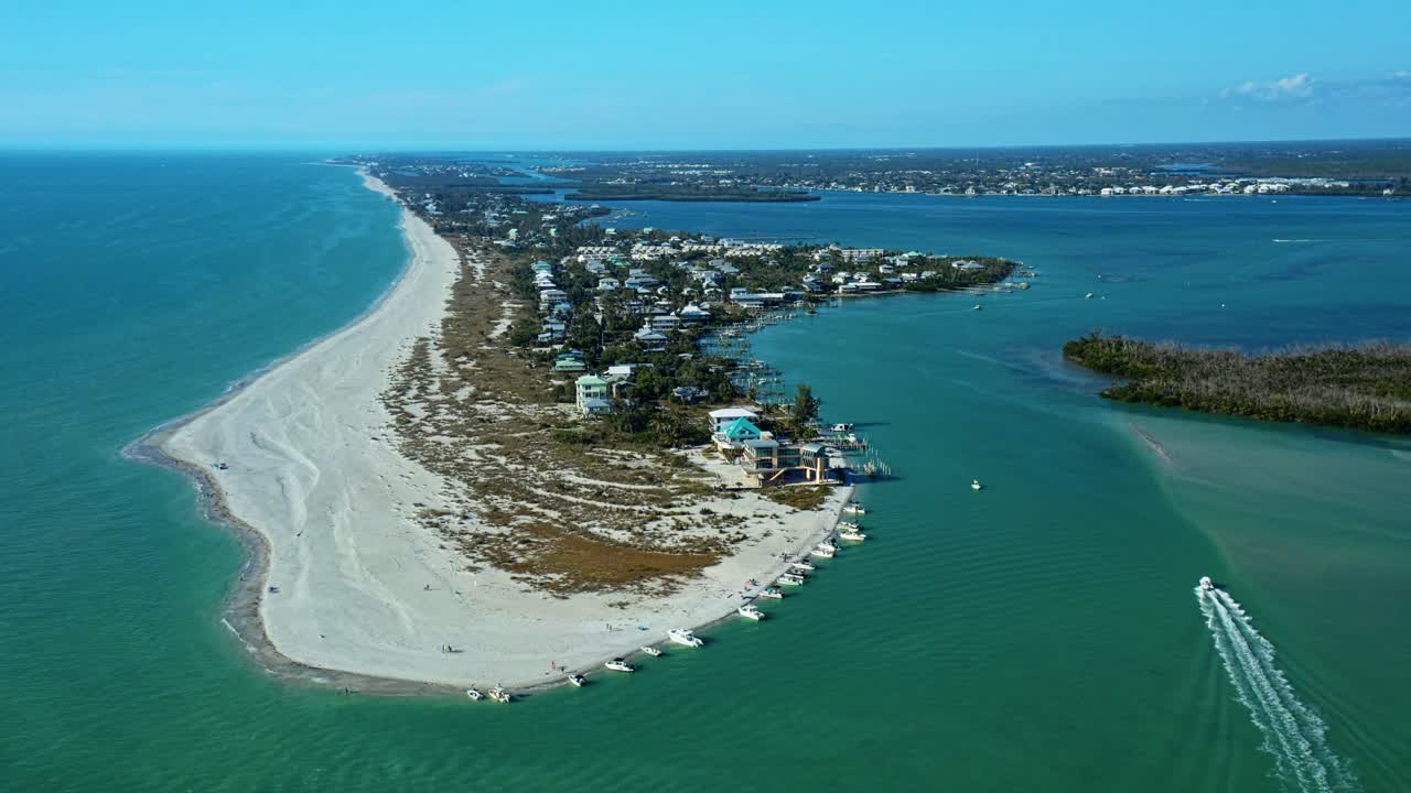 Kysthalvø med sandstrande og beboelseshuse, med både for anker nær kysten i et roligt bugtområde under klar blå himmel på en solrig dag.