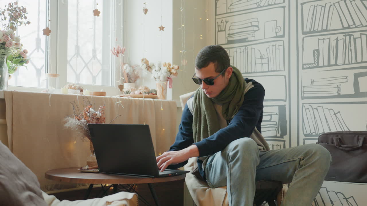 Caucasian man typing on laptop, focused freelancer working in cozy corner, sunlight through window, scarf and sunglasses, coffee mug and notepad on table, modern remote workspace, productive digital