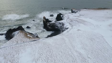 Aerial-orbiting-shot-of-snowy-coastline-and-waves-of-ocean-against-basalt-cliffs---Gatklettur,Iceland