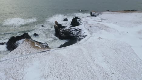 aerial orbiting shot of snowy coastline and waves of ocean against basalt cliffs - gatklettur,iceland