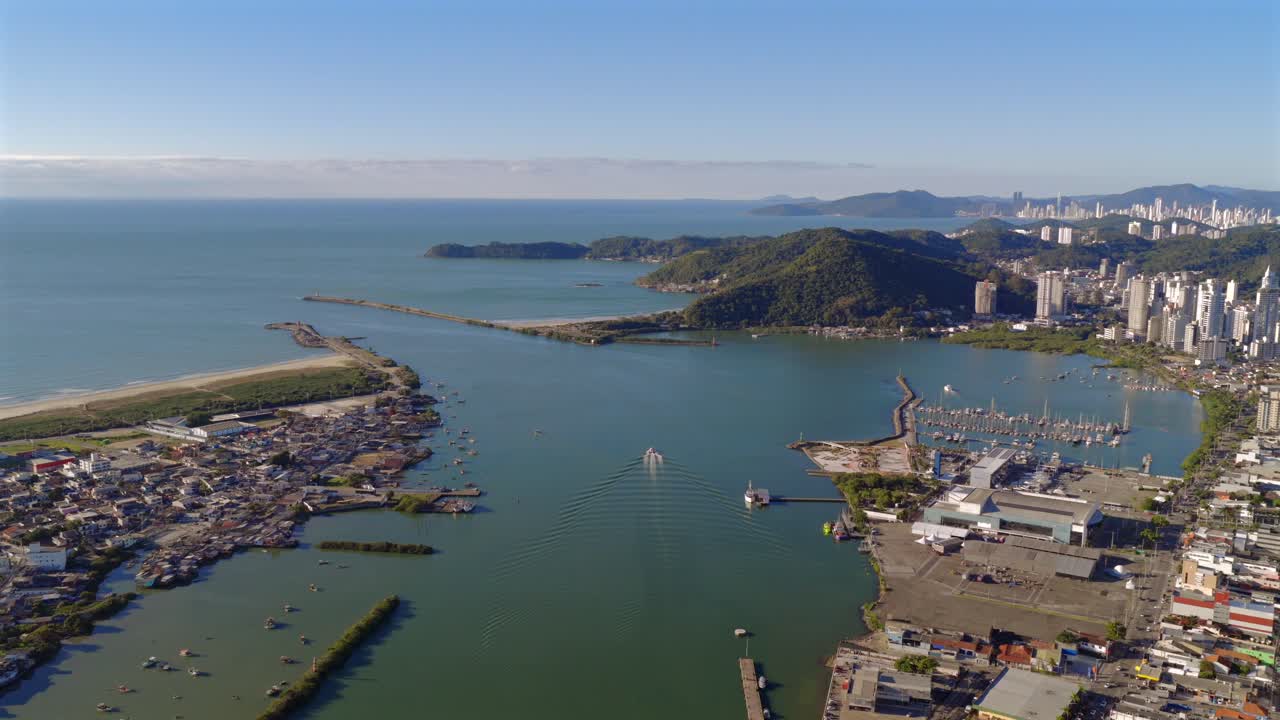 Drone over Itajaí river to ocean horizon, city beneath, clear afternoon skies. Boat on water