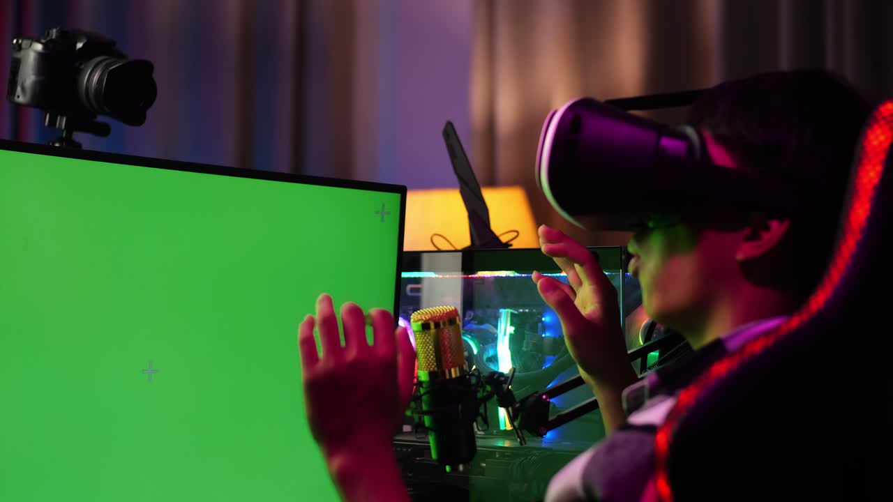 Close Up Back View Of Asian Boy Streamer Playing Game, Touching, And Swiping Experience Virtual Reality In Futuristic Goggles. Green Screen Personal Computer On Desk Illuminated By Rgb Led Strip Light