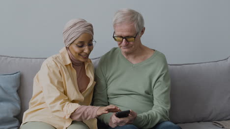sonriente mujer árabe de mediana edad ayudando a un anciano a usar un smartphone mientras se sienta en un sofá en casa