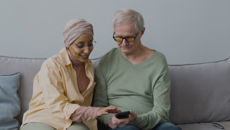 smiling middle aged arabic woman helping a senior man to use smartphone while sitting on sofa at home