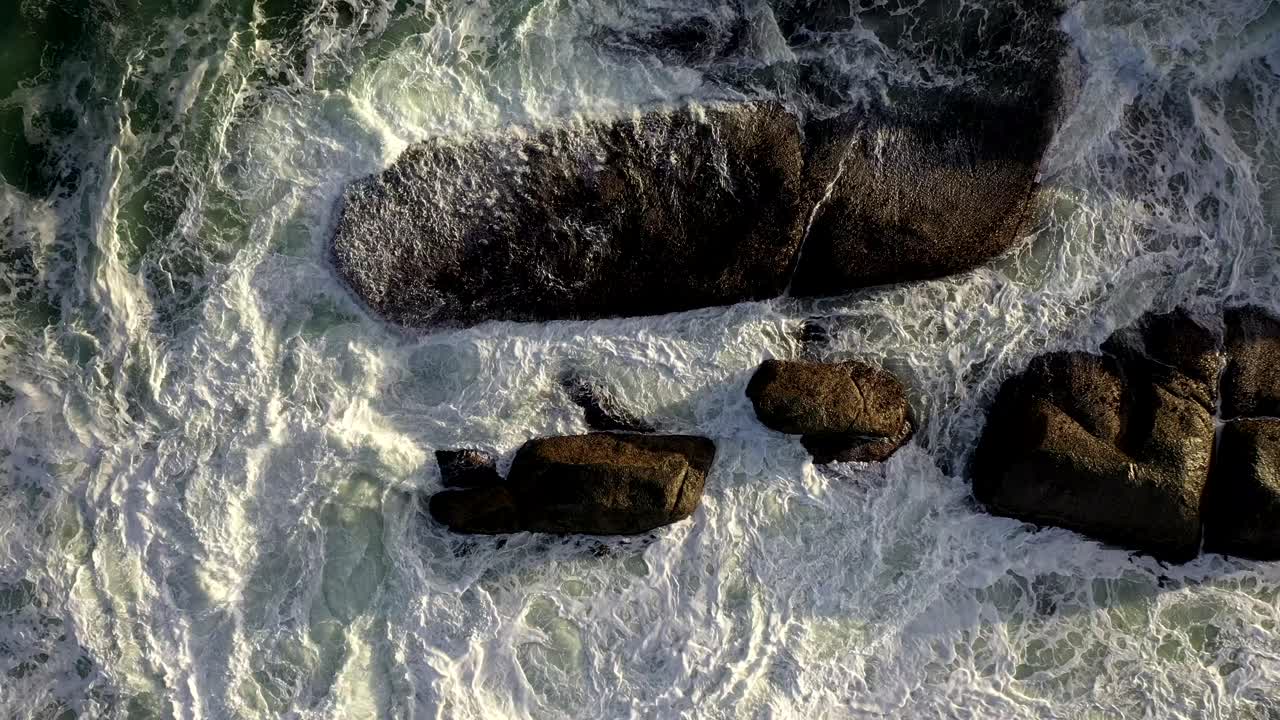 Splendida ripresa cinematografica cenitale in lento sollevamento di onde oceaniche agitate che si infrangono contro le rocce durante il tramonto a Camps Bay Beach, Città del Capo.
