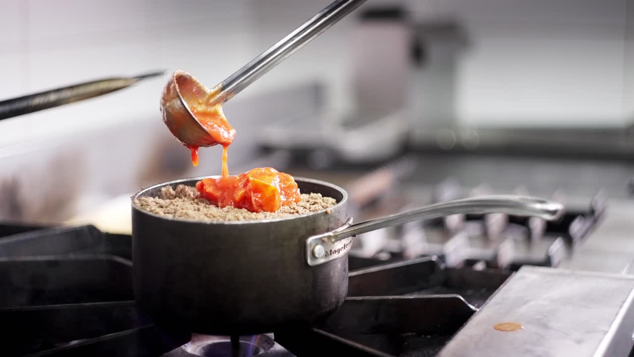 Close up view in slow motion of a Cooking spoon pouring tomato sauce over ground beef in a restaurant kitchen.