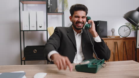 Indian-smiling-businessman-talking-on-wired-vintage-telephone-of-80s,-says-hey-you-call-me-back