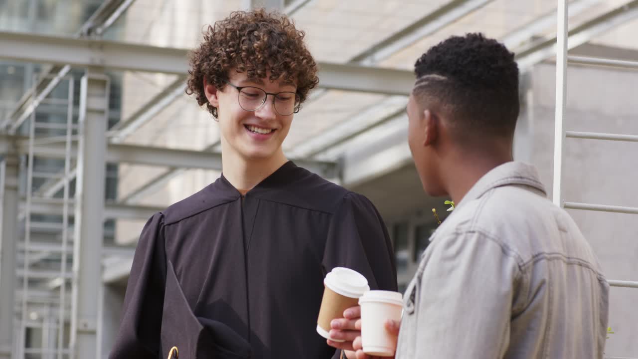 Diverse male friends celebrating graduation in robe and cap handing coffee and sipping on campus