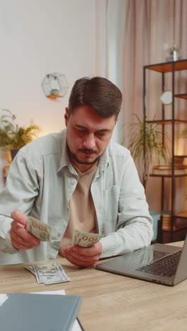rich happy caucasian man counting money cash using laptop calculate domestic bills at home table