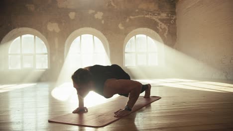 A-male-athlete-in-a-black-sports-summer-uniform-does-push-ups-on-a-special-mat-and-lifts-one-of-his-legs-during-push-ups.-Unique-push-up-technique.-Sports-activities-in-a-sunny-brick-hall