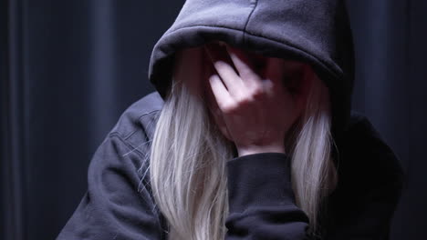 chica rubia asustada con capucha negra sosteniendo su cabeza cubriendo su rostro, luciendo estresada, deprimida en una habitación con poca luz