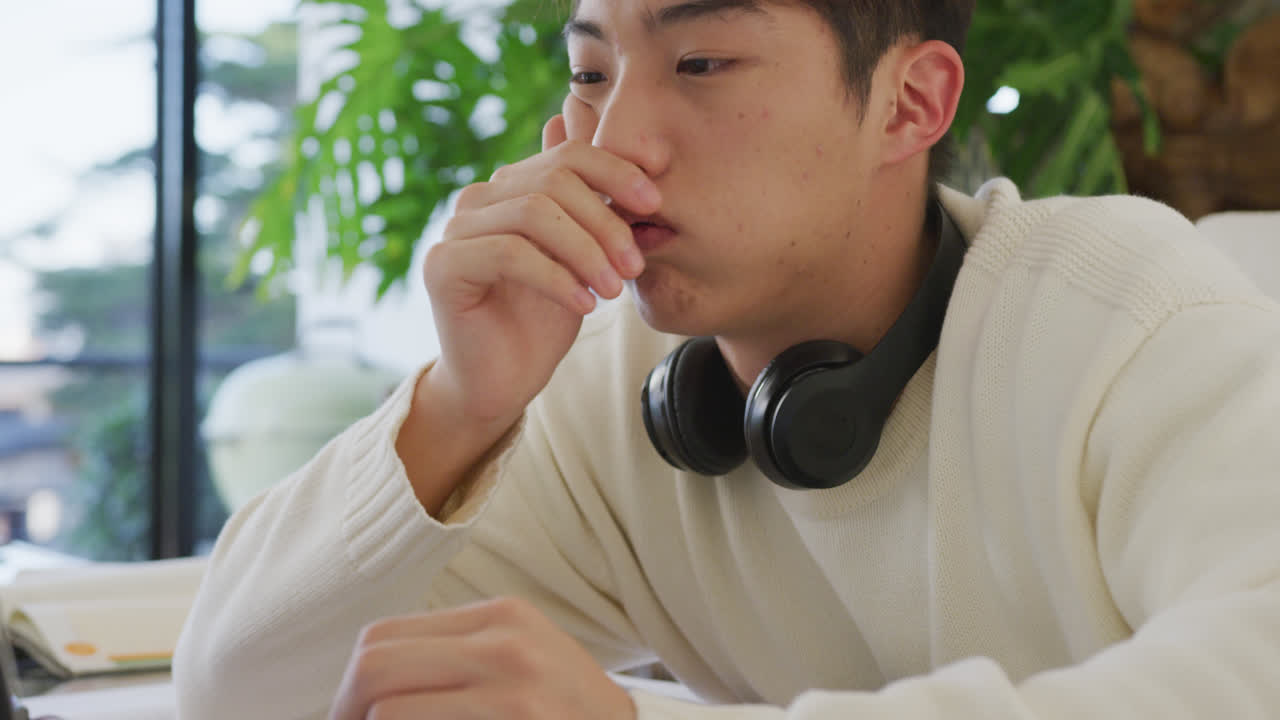 헤드폰을 착용한 아시아 남성 십대 청소년이 거실에서 노트북을 배우고 사용하고 있습니다.