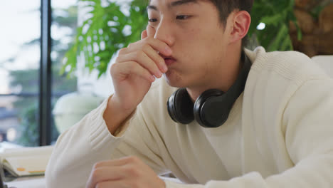 헤드폰을 착용한 아시아 남성 십대 청소년이 거실에서 노트북을 배우고 사용하고 있습니다.