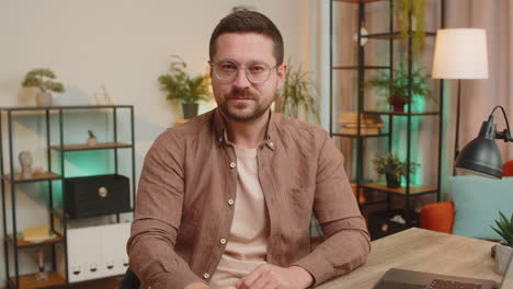 caucasian young businessman presses index finger to lips makes silence gesture at home office table