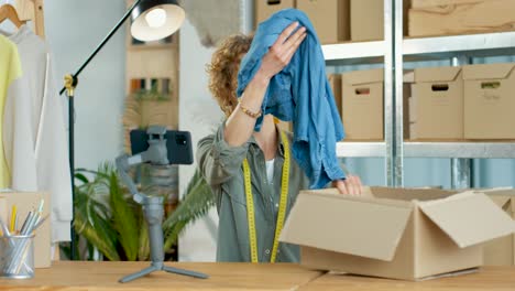 Mujer-Joven-Caucásica-Hablando-Grabando-Video-Para-Un-Blog-De-Moda-En-Una-Cámara-De-Teléfono-Inteligente-Sentada-En-Su-Propio-Estudio-De-Tienda-De-Ropa-Desempacando-Un-Paquete-Que-Muestra-Ropa,-Concepto-De-Diseñador